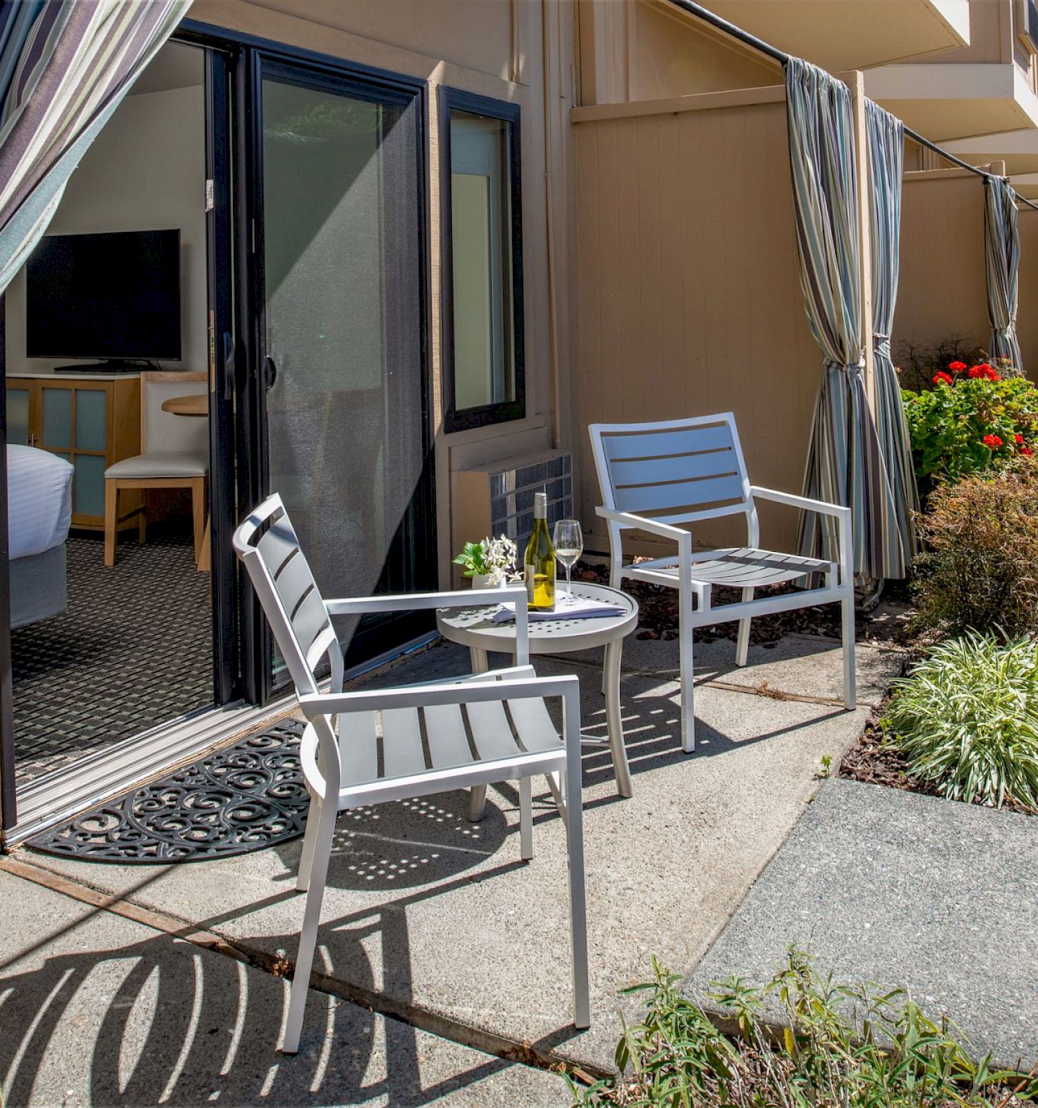 Outdoor patio with two chairs, a small table with drinks, and an open sliding door leading inside. Striped curtains on either side.