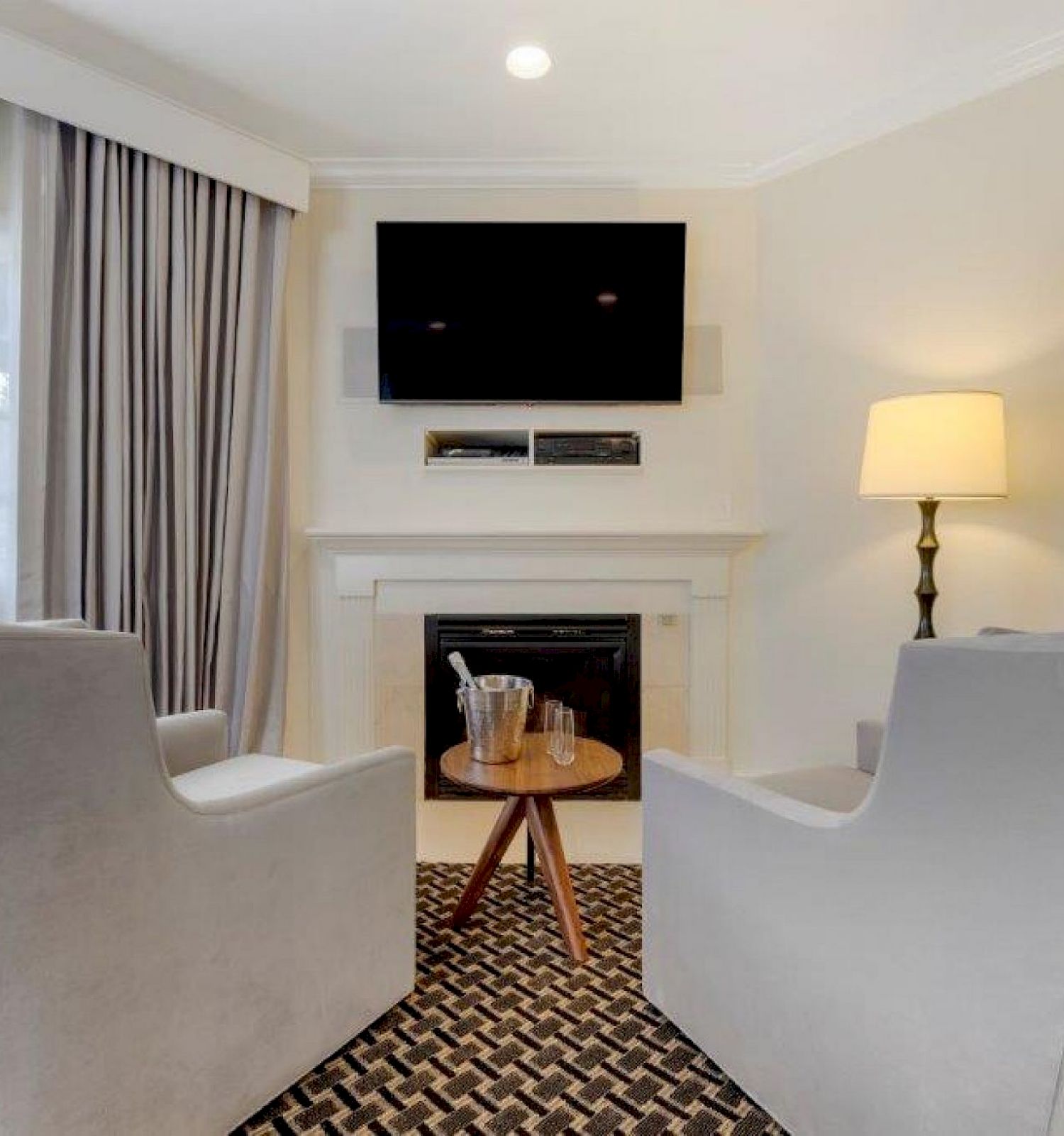 A modern living room features two grey armchairs, a small round table, a lamp, a TV mounted above a fireplace, and large windows with curtains.