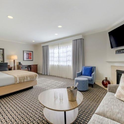 A modern, spacious hotel room with a bed, work desk, sofa, round table, and TV mounted above a fireplace. The window is draped with curtains.