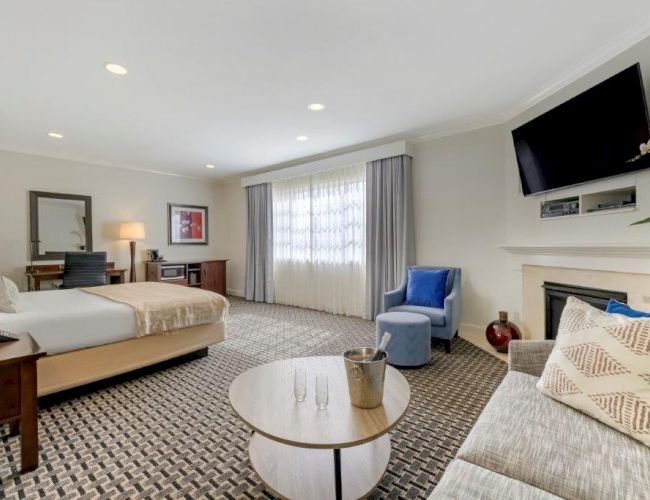 A modern, spacious hotel room with a bed, work desk, sofa, round table, and TV mounted above a fireplace. The window is draped with curtains.