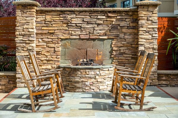 Four wooden rocking chairs surround an outdoor stone fireplace on a patio.