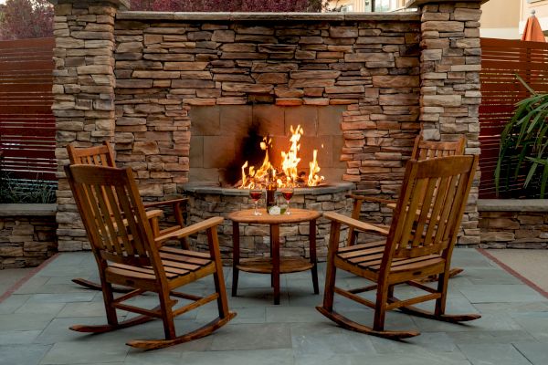 Two wooden rocking chairs face a lit outdoor firepit, with a small table and drinks between them, set against a stone wall backdrop.