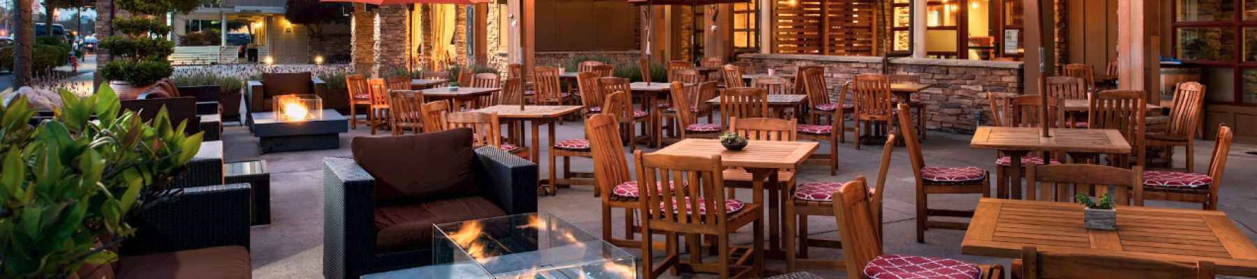 Outdoor patio with wooden tables, red umbrellas, string lights, and fire pits, surrounded by greenery and seating under a clear sky.