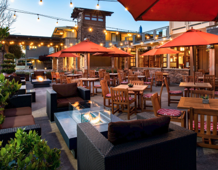 Outdoor patio with wooden tables, red umbrellas, string lights, and fire pits, surrounded by greenery and seating under a clear sky.