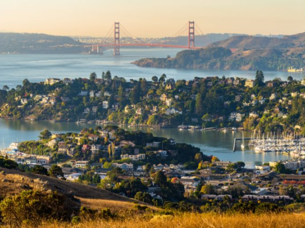 A scenic coastal town nestled on hills by winding water, with a marina, houses, and boats, plus the Golden Gate Bridge in the distance.