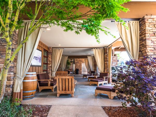 A cozy outdoor lounge under beige drapes with wooden chairs, a stone pillar entry, green trees, and a spa-like seating area in warm ambiance.