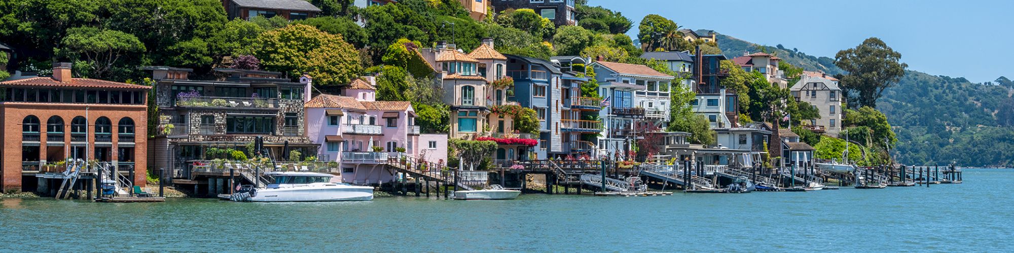 A row of colorful houses perched on a hillside along a calm waterfront, with boats docked by the shore and blue sky above.