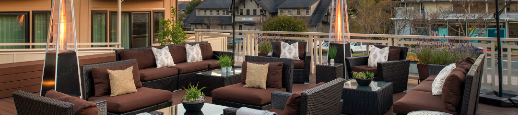 A stylish outdoor lounge on a wooden deck with wicker sofas, blue umbrellas, tall patio heaters, and a scenic hillside view, perfect for socializing.