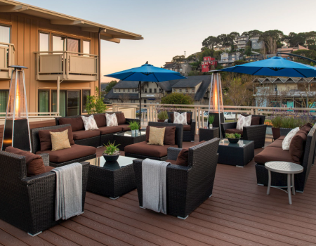 A stylish outdoor lounge on a wooden deck with wicker sofas, blue umbrellas, tall patio heaters, and a scenic hillside view, perfect for socializing.