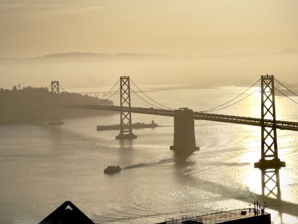 A suspension bridge spans misty water at sunrise, with cables, piers, and a ship cruising below.