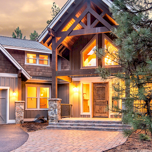 The image shows a modern, well-lit house surrounded by trees, featuring a three-car garage, wooden exterior, large windows, and a stone walkway ending the sentence.