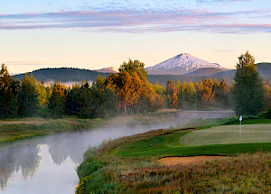 A serene landscape featuring a golf course by a river, with mist rising and a snow-capped mountain in the background, surrounded by lush trees.
