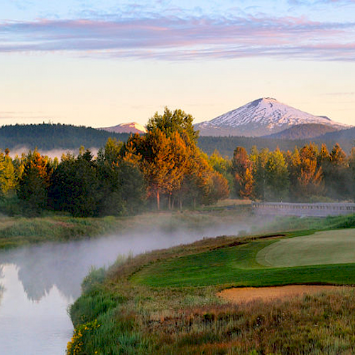 A serene landscape featuring a golf course by a river, with mist rising and a snow-capped mountain in the background, surrounded by lush trees.