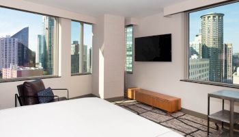 A modern hotel room with large windows offering a city view, a wall-mounted TV, a chair, and a simple wooden bench.