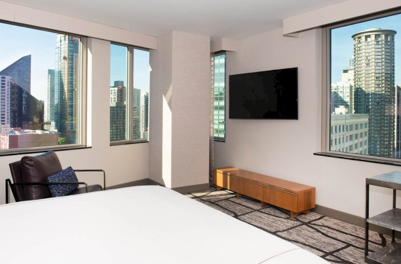 A modern hotel room with large windows offering a city view, a wall-mounted TV, a chair, and a simple wooden bench.