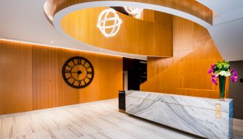 The image shows a modern reception area with a marble desk, a large wall clock, wooden accents, and elegant light fixtures.