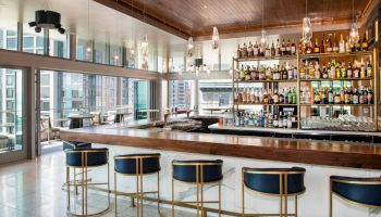 The image shows a modern, stylish bar with a wooden countertop, lined with blue barstools, and a large selection of bottles on shelves behind it.