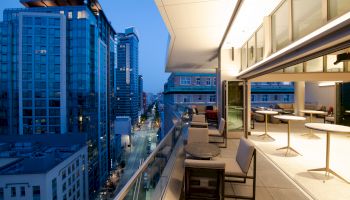 A modern balcony with seating overlooks a city street lined with tall, illuminated buildings during twilight, capturing an urban, sophisticated vibe.