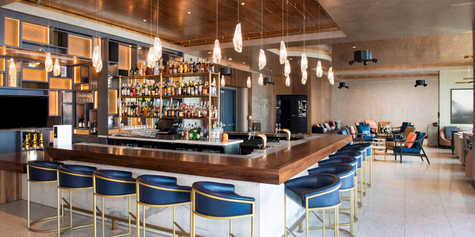 A modern bar with a wooden countertop, surrounded by blue barstools, a well-stocked liquor shelf, hanging lights, and a seating area in the background.