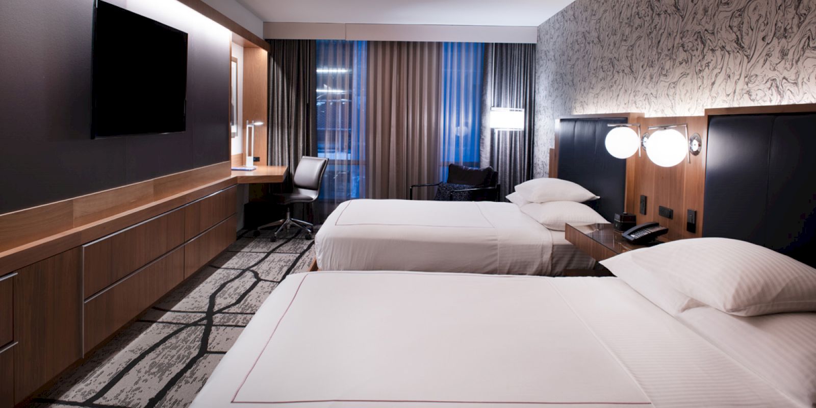 A modern hotel room with two neatly made beds, a wall-mounted TV, a work desk with a chair, and large windows with curtains ends the sentence.