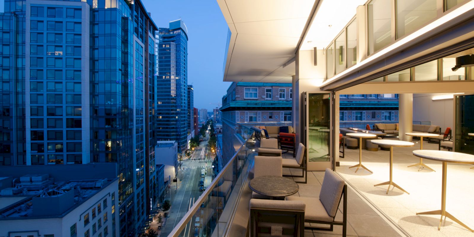 A modern balcony with seating and tables overlooking a bustling cityscape with tall buildings, captured during the early evening.