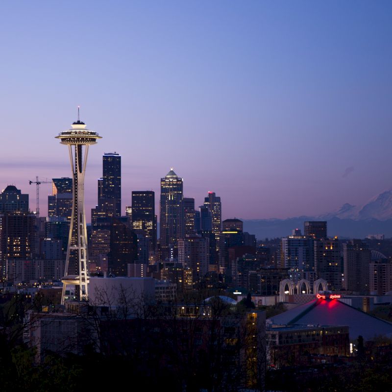 A cityscape of Seattle at dusk, featuring the iconic Space Needle, high-rise buildings, and Mount Rainier in the background.