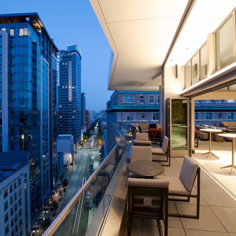 A modern cityscape at dusk viewed from a balcony with outdoor seating, tables, and a view of surrounding high-rise buildings.