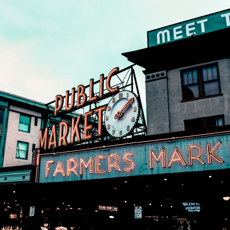 The image shows a public market with a clock and neon signs reading "Public Market" and "Farmers Market" on the facade of two buildings.