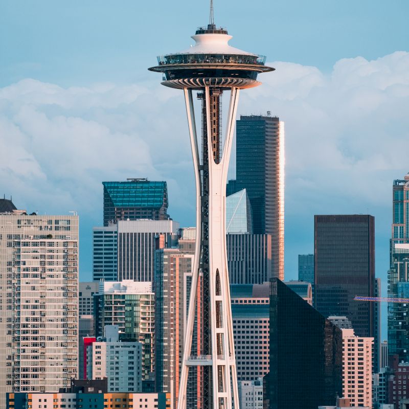 The image features the Space Needle with a backdrop of Seattle's skyline, showing various modern buildings and skyscrapers under a partly cloudy sky.