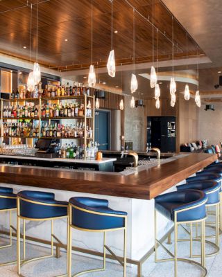 The image shows a modern bar with a wooden counter, blue cushioned stools, hanging light fixtures, and shelves stocked with various bottles.