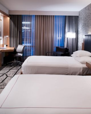 A modern hotel room with two neatly made beds, a wall-mounted TV, a work desk with a chair, lamps by the beds, and large windows with curtains.