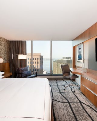 A modern hotel room with a large bed, a flat-screen TV, a desk with a chair, and a view of tall buildings through the window.