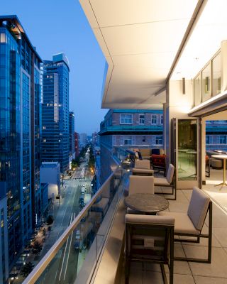 A modern cityscape at dusk with high-rise buildings lined up; a spacious balcony with tables and chairs, large windows, and bright indoor lighting.