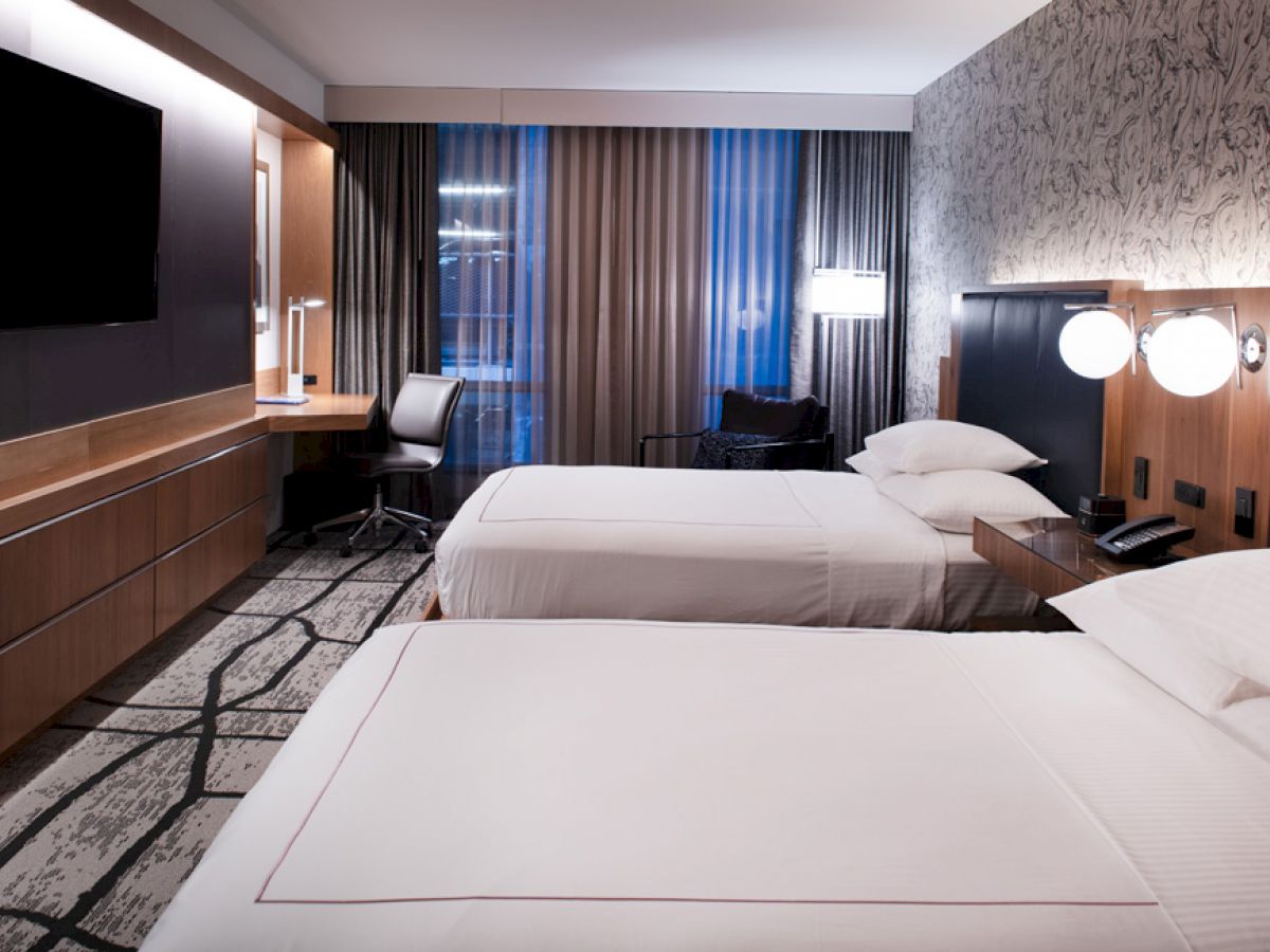 A modern hotel room with two beds, a desk with a chair, a wall-mounted TV, and stylish decor featuring a patterned wall and carpet.