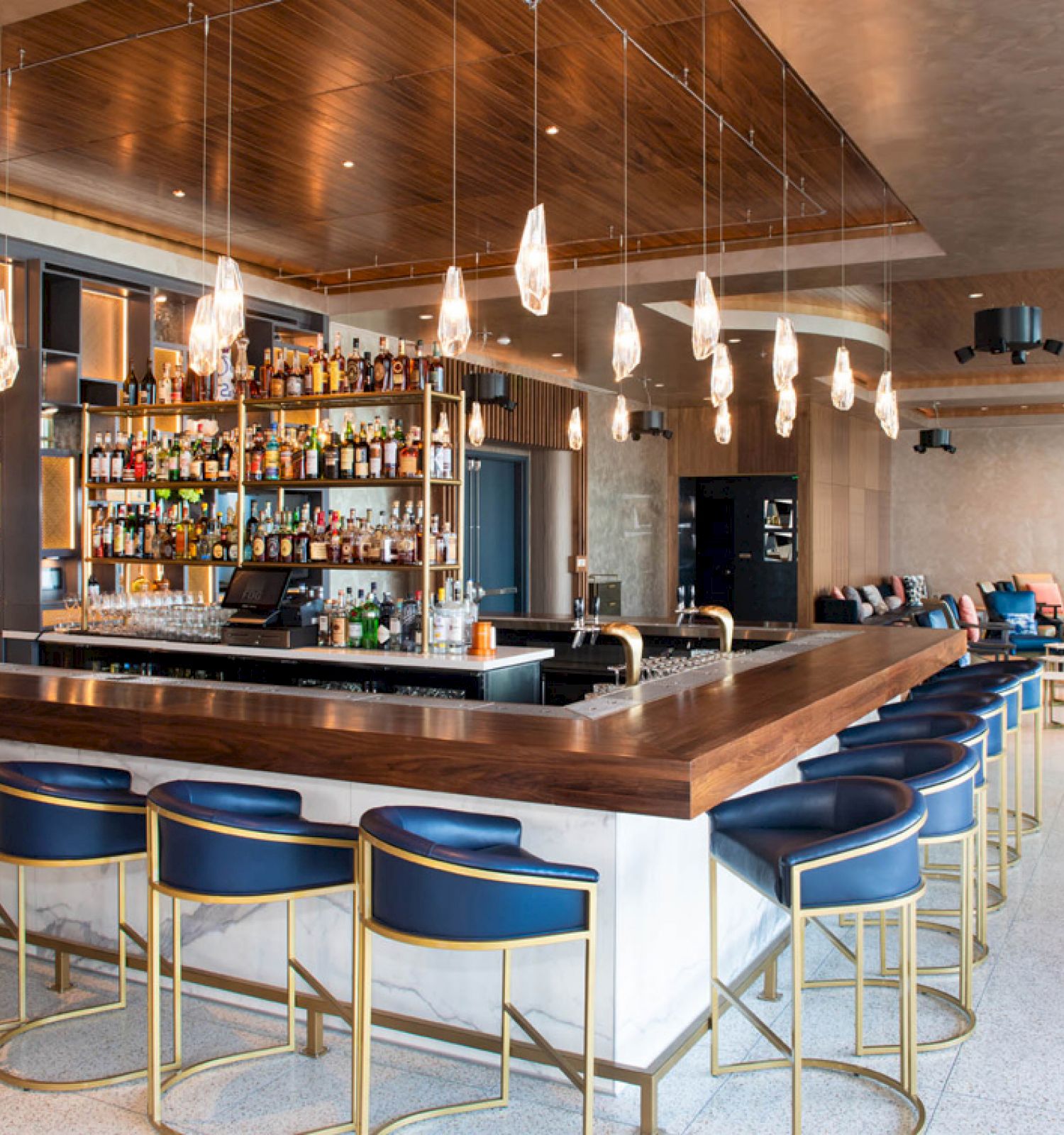 A modern bar with a wooden counter, blue and gold stools, hanging pendant lights, and well-stocked shelves of liquor bottles. The bar is spacious.