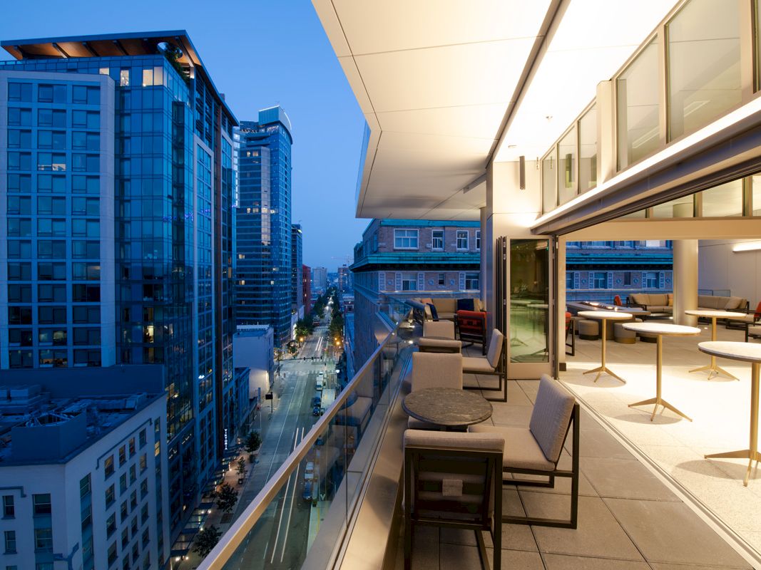The image shows a modern balcony with city views at dusk, featuring lounge chairs, tables, and skyscrapers in the background.