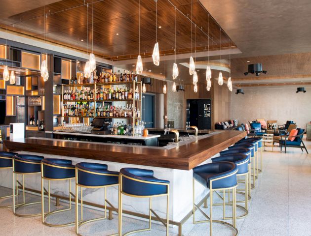 A modern bar with a wooden countertop, illuminated by hanging lights, and surrounded by blue stools. A variety of bottles are displayed behind the bar.