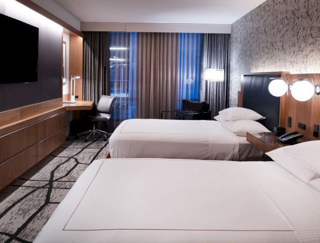 A modern hotel room with two neatly made beds, a flat-screen TV on the wall, a work desk with a chair, and ambient lighting.