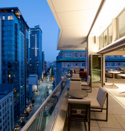 A balcony overlooking a cityscape with modern buildings at dusk; outdoor seating and tables extend from an indoor area.