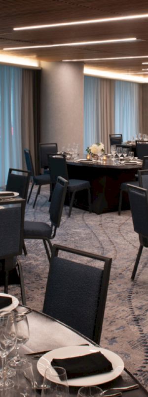 A modern dining room setup with round tables, black chairs, and place settings. The space is elegantly lit, with large windows featuring blue curtains.