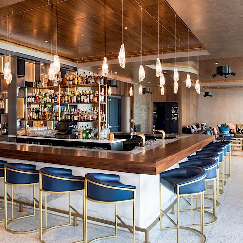 The image shows a modern bar with a large selection of drinks, elegant bar stools, hanging lights, and a cozy seating area in the background.
