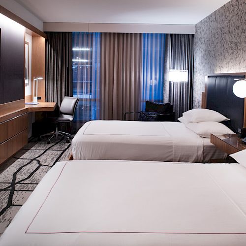 A modern hotel room with two neatly made beds, a wall-mounted TV, a desk with a chair, stylish lighting, and contemporary decor ending the sentence.