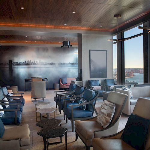 This image shows a modern lounge area with comfortable seating, large windows offering a waterfront view, and a mural on the far wall ending the sentence.