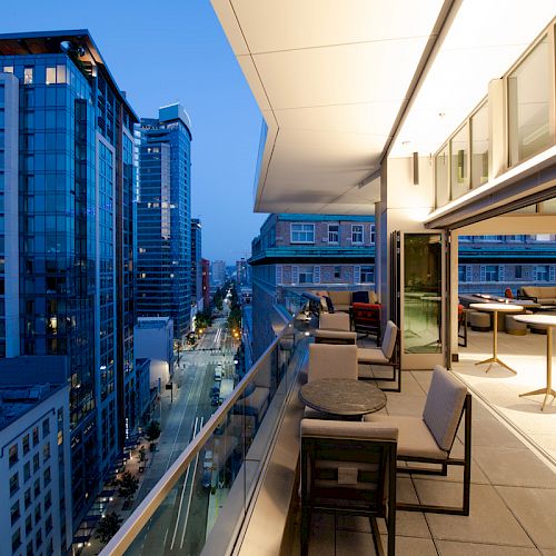 A modern cityscape at dusk, featuring tall buildings and a stylish roof terrace with tables, chairs, and large windows overlooking the view.