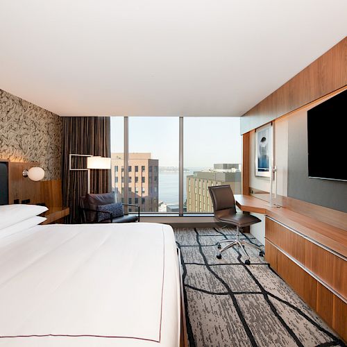 A modern hotel room with a large bed, flat-screen TV, desk, and chair, featuring floor-to-ceiling windows with a city view in the background.