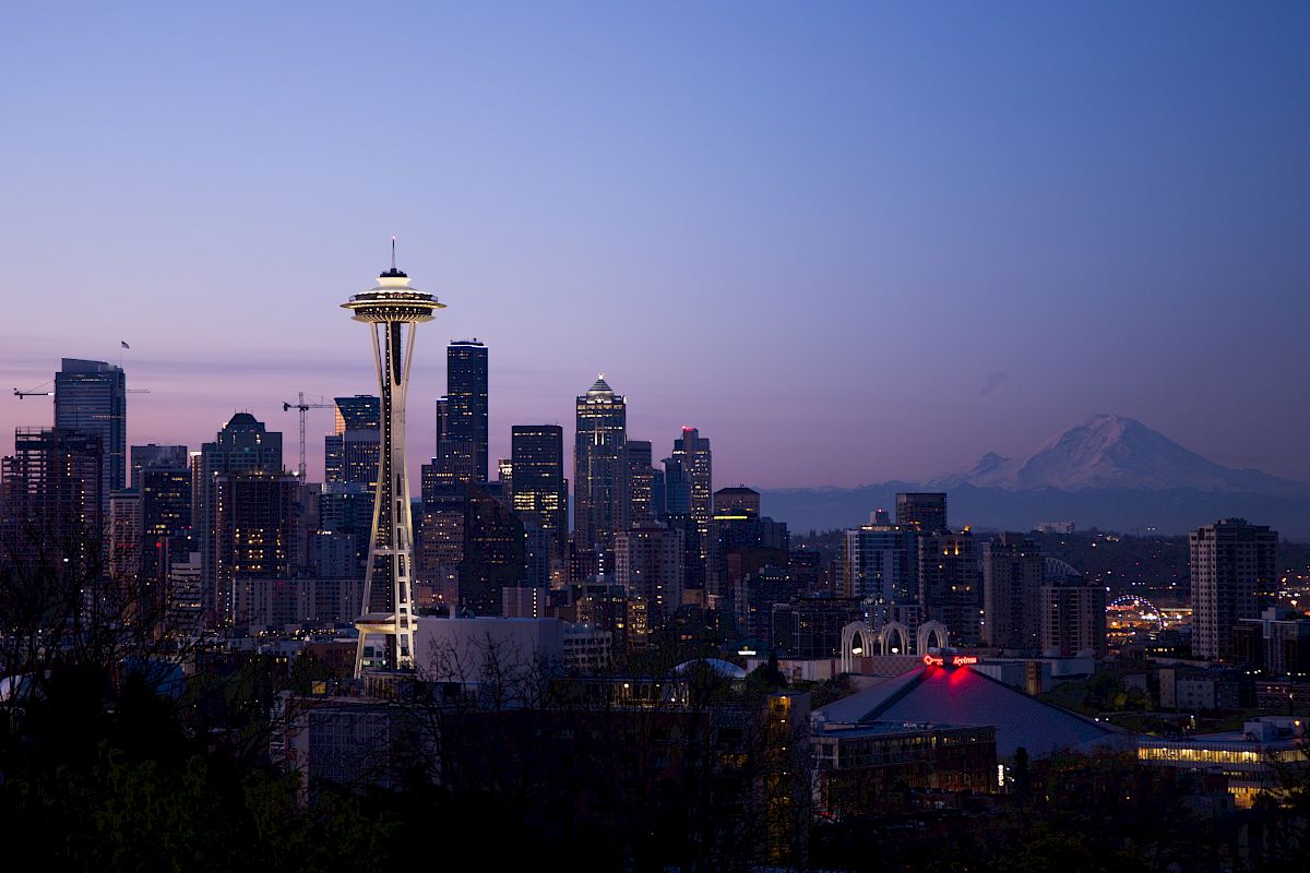 The image shows Seattle's skyline during sunset, featuring the iconic Space Needle and Mount Rainier in the background, framed by modern skyscrapers.