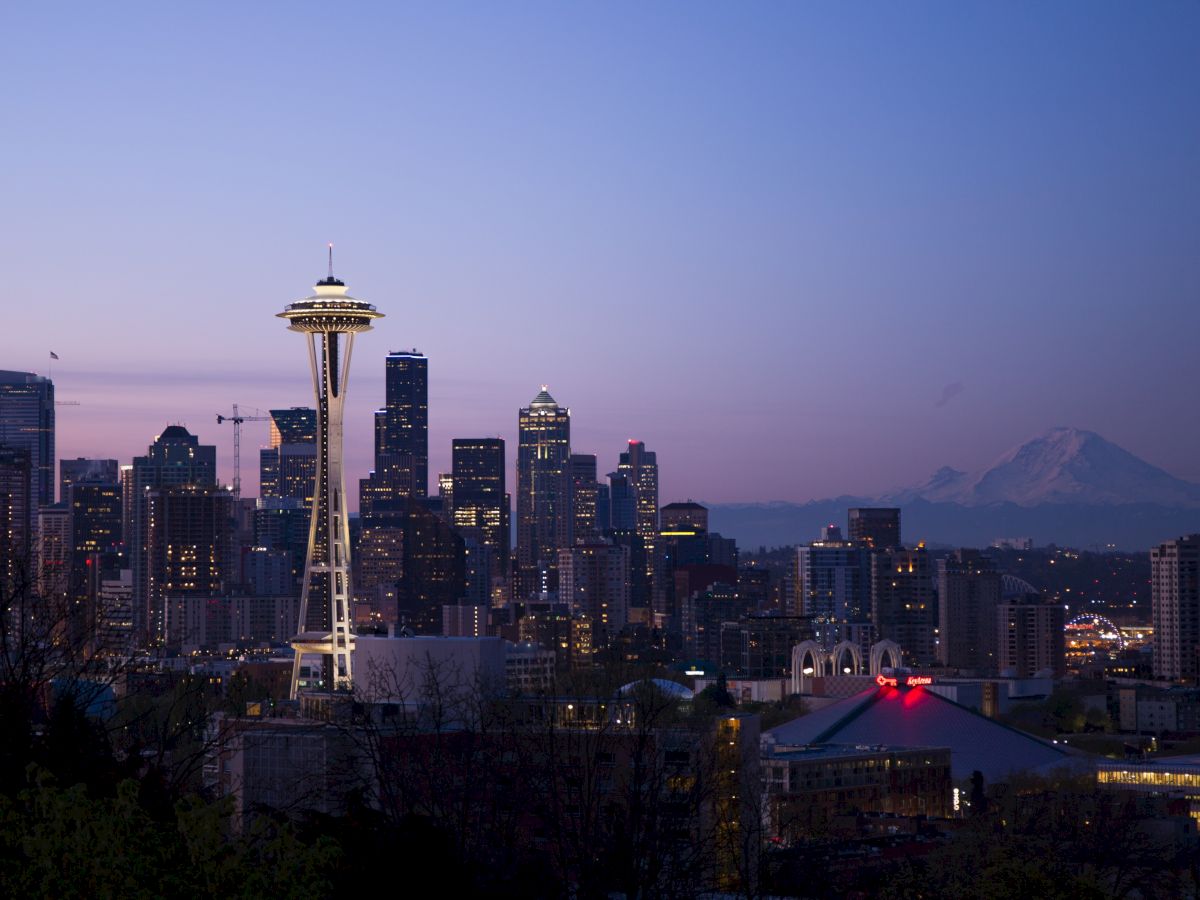 The image shows Seattle's skyline during sunset, featuring the iconic Space Needle and Mount Rainier in the background, framed by modern skyscrapers.