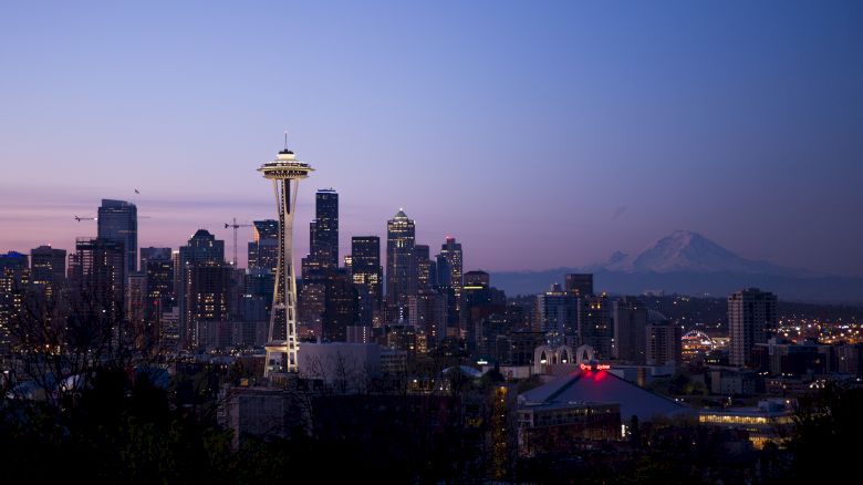 The image shows Seattle's skyline during sunset, featuring the iconic Space Needle and Mount Rainier in the background, framed by modern skyscrapers.