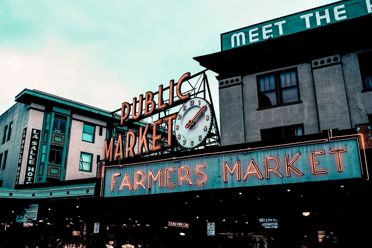 The image displays a street view of a "PUBLIC MARKET" sign with an adjacent "FARMERS MARKET" neon sign and surrounding buildings in an urban area.