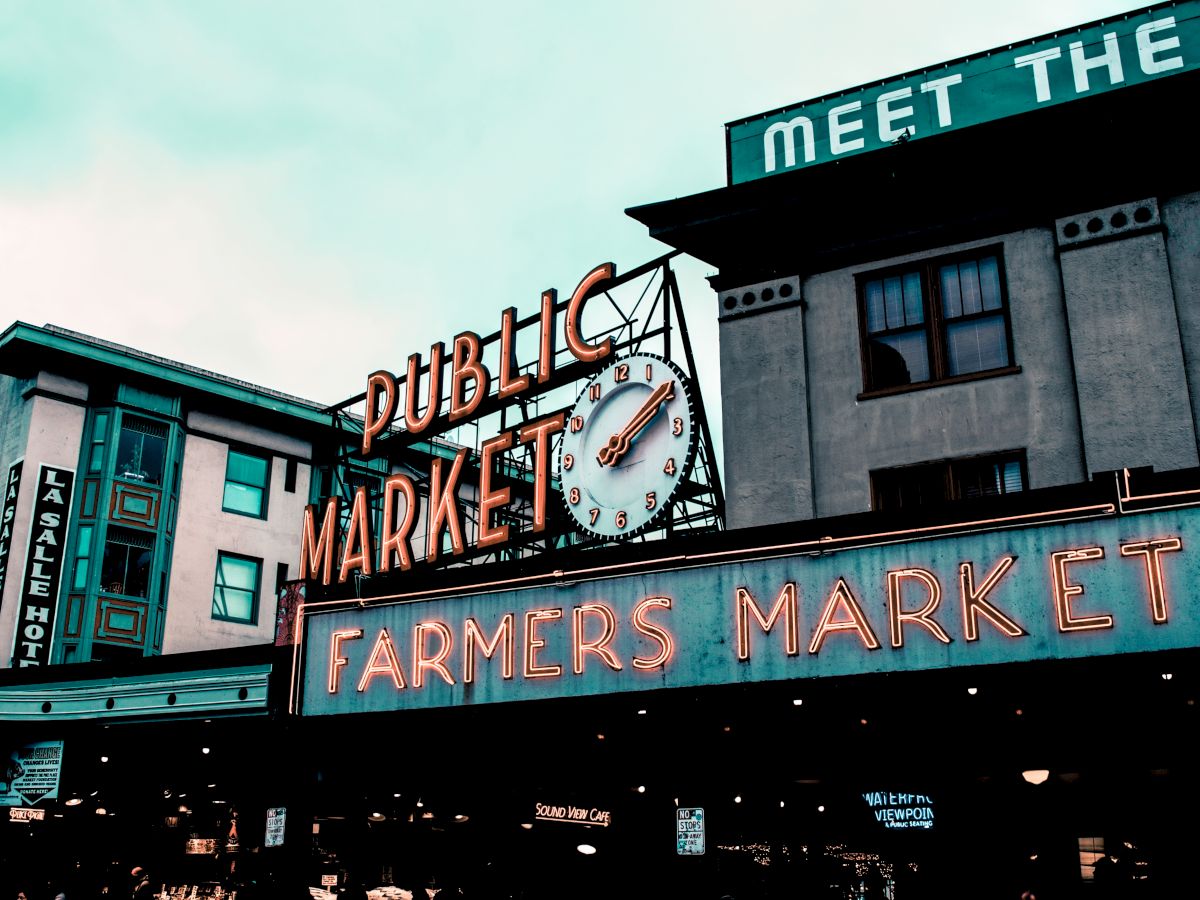 The image displays a street view of a "PUBLIC MARKET" sign with an adjacent "FARMERS MARKET" neon sign and surrounding buildings in an urban area.